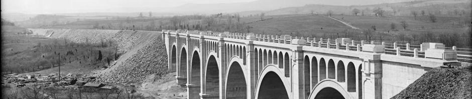Black and white photo of bridge shortly after completion with bare hills leading from the river below to the top of the bridge abutments.