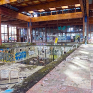 Facing hallway windows from other end of indoor pool room with view of graffitied, empty pool